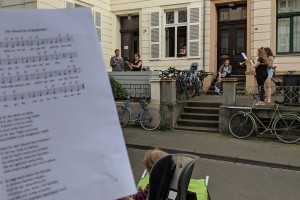 Flyer mit Noten und Liedtext, dahinter einige Sänger*innen vor einem Wohnhaus