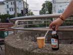 Ein Pappbecher mit Strohhalm und eine Falsche Bier stehen auf einem Betonmäuerchen. Eine Hand greift nach der Flasche.