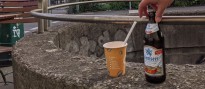Ein Pappbecher mit Strohhalm und eine Falsche Bier stehen auf einem Betonmäuerchen. Eine Hand greift nach der Flasche.