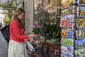 Eine Frau steht vor einem Blumenladen und hält eine Topfpflanze in der Hand. Auf einer Bank vor dem Schaufenster stehen Blumen und eine Geldkassette.