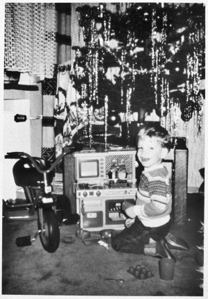 Ein Junge spielt mit einer Spielzeugküche und lacht in die Kamera. Um ihn herum sind Küchenutensilien verteilt, links von der Spielzeugküche steht ein Dreirad. Hinter der Spielzeugküche ist ein Weihnachtsbaum aufgebaut, an dem Lametta hängt.