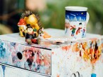 Auf einem Schreibtisch mit Büroutensilien wie Hefter, Block und Stifthalter steht ein Schubladencontainer aus bunter Pappe. Darauf diverse Kleinigkeiten: Eine Tasse mit winterlichem Motiv, ein Miniaturflugzeug, ein Keramikigel und eine Schornsteinfegerfigur. 