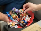 Eine Hand hält einen Süßigkeitenteller über einer geöffneten Schublade mit einer Aktenmappe. Eine andere Hand greift nach einer Praline. Auf dem Teller befinden sich zum Beispiel ein Schokoladen-Weihnachtsmann und ein Osterhase aus Schokolade.