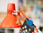 An einer roten Schreibtischlampe ist mit einem Magnet eine Weihnachtskarte befestigt. Auf der Karte steht: „Weihnachtsmarkt im Stadtgarten“, das Motiv darunter zeigt viele bunte Weihnachtskugeln.