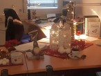 Durch eine geöffnete Tür fällt der Blick auf eine Schreibtischlandschaft. Große Teile sind mit diversen weihnachtlichen Objekten bedeckt: Dosen, Tischdecken, Figuren. Auf einem anderen Teil rund um den PC liegen Unterlagen. Im Hintergrund leuchtet eine Lichterkette. 