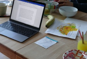 Aufgeklappter Laptop auf einem Tisch neben Tassen, Schalen, einer Banane, Pinseln im Wasserglas und einem gemalten Regenbogenbild "Alles wird gut".