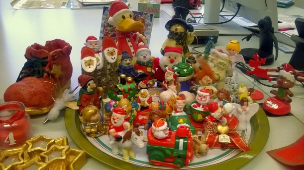 Auf einem Schreibtisch steht ein Teller mit diversen zum Teil sehr bunten Weihnachtsobjekten. Darunter Engel, Weihnachtsmänner, Rentiere. Um den Teller stehen Deko-Objekte wie beispielsweise ein roter Nikolausstiefel aus Keramik.