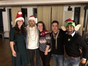 Fünf Personen, zwei Frauen und drei Männer stehen nebeneinander und haben die Arme um die Schulter der Nebenstehenden gelegt. Sie tragen zum Teil Weihnachtsmützen auf dem Kopf und lange silberfarbene Weihnachtsbaumketten um den Hals.