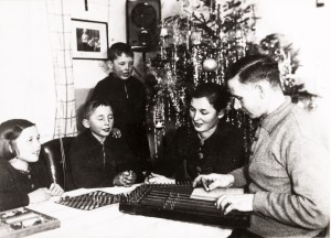 An einem Tisch vor einem Weichnachtsbaum sitzen ein Mädchen, ein Junge, einer Frau und ein Mann. Der Mann spielt auf der Zither, die Kinder spielen ein Brettspiel. Ein weiterer Junge steht vor dem Christbaum,