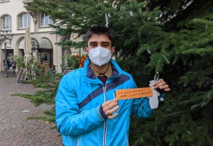 Ein junger Mann mit FFP2-Maske steht vor einem Weihnachtsbaum und hält ein Schild in die Kamera: „Ich wünsche mir mehr Gemeinschaft“. Das Schild hängt an dem Baum.