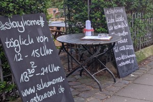 Foto aus der Zeit eines Corona-Lockdowns, auf dem ein Restaurant mit zwei Tafeln seinen Außerhaus-Verkauf bewirbt. Als Überschrift auf den Tafeln wurde „#stayhome“ gewählt.