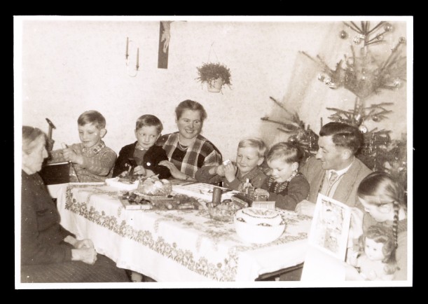 Schwarz-weiß Foto zeigt eine Familie am weihnachtlich dekorierten Tisch, neben dem ein Weihnachtsbaum steht. Die Kinder halten Geschenke in den Händen.