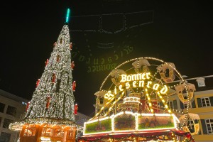 Nächtliche Ansicht des Bonner Weihnachtsmarktes. Lichterketten beleuchten eine Glühweinbude, Fahrgeschäfte und einen großen Weihnachtsbaum. Ein nostalgisches Riesenrad ziert die Leuchtreklame: „Bonner Riesenrad“.