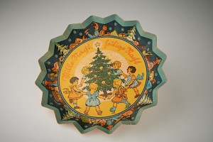 Pappteller mit Wellenrand und weihnachtlichem Bildmotiv: Sechs Kinder tanzen um einen geschmückten Tannenbaum und singen. Über dem Bild steht in deutscher Schrift der Titel des Weihnachtsliedes: "Stille Nacht – Heilige Nacht".