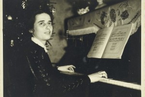 Eine Frau spielt an einem Klavier ein Musikstück. Vor ihr stehen die Noten aufgeschlagen. Im Hintergrund ist ein geschmückter Weihnachtsbaum zu sehen.
