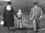 Eine Frau und ein Mann gehen mit einem Jungen in ihrer Mitte, der ein Fernglas um den Hals hängen hat auf einer Wiese spazieren, im Hintergrund sind Hügel erkennbar. 