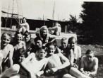 Jungen in Badehosen, die auf der Wiese liegen und sitzen, posieren zu einem Gruppenfoto. Einer hat ein Handtuch über den Kopf gelegt. Im Hintergrund sind ein Schwimmbecken und Sprungbretter zu erkennen. 