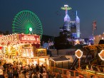 Beleuchtete Fahrgeschäfte auf einer Kirmes mit vielen Besucher*innen im Vordergrund. Zu erkennen sind unter anderem eine Achterbahn und ein Riesenrad