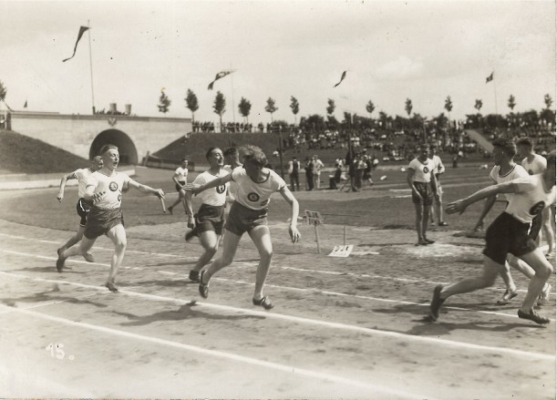 Eine Schwarz-weiß-Aufnahme zeigt die Laufbahn eines Stadions und Staffelläufer bei der Staffelstabübergabe.