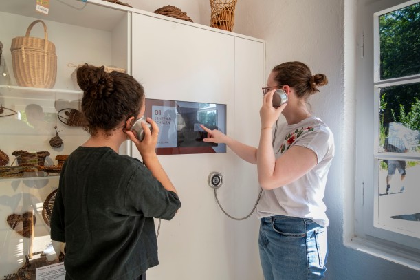 Zwei junge Menschen stehen an einer Medienstation im Museum, berühren einen Touch Screen und haben Kopfhörer am Ohr.