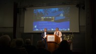 Das Foto zeigt zwei Personen auf einem Podium. Im Hintergrund ist eine Leinwand zu sehen, auf der das Foto einer Kirche erscheint und geschrieben steht: Filmpremiere. Zwischen Heimat und Kulturerbe. Abschied von der evangelischen Versöhnungskirche in Overath. Schemenhaft sind die Köpfe von Zuschauer*innen zu erkennen. Eine Bildunterschrift sagt: Die Tagung eröffnete mit einer feierlichen Filmpremiere.