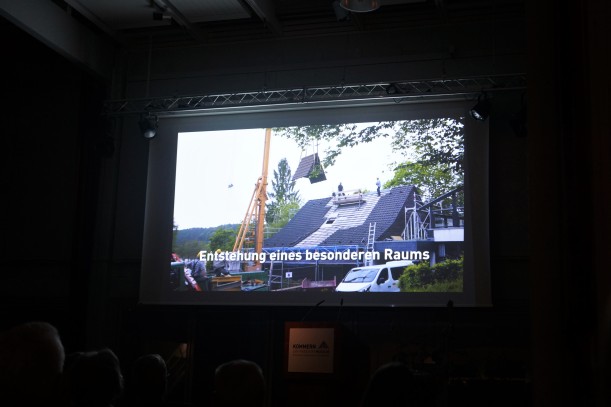 Das Foto zeigt eine Leinwand, auf der ein Dokumentarfilm abgespielt wird. Zu sehen ist ein Haus, dessen Dach abmontiert wird. Im Untertitel steht: Entstehung eines besonderen Raums. Schemenhaft sind die Köpfe von Zuschauer*innen zu erkennen.