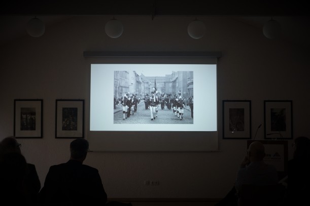 Das Foto zeigt eine Leinwand, auf der ein Dokumentarfilm abgespielt wird. Zu sehen ist eine schwarz-weiße Fotografie von Männern in Uniform. Eine Bildunterschrift sagt: Auf dem Programm standen Filmvorführungen ...