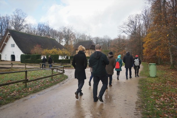 An zwei alten Fachwerkhäusern vorbei laufen in Jacken, Mützen und Schals gekleidete Personen durch eine herbstliche Landschaft.