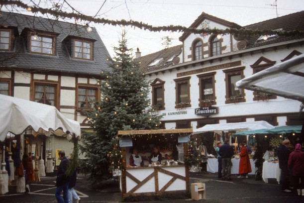 Weihnachtsmarkt mit mehreren weihnachtlich geschmückten Verkaufsständen und einem Tannenbaum mit Lichterschmuck in der Mitte. Im Vordergrund steht eine Holzhütte, die mit Lichterkette sowie links und rechts einem kleinen, geschmückten Tannenbaum dekoriert ist. In der Hütte stehen zwei Frauen mit weißen Schürzen, die wahrscheinlich Weihnachtsgebäck verkaufen. Hinter dem Stand ist ein großer, mit Lichterkette geschmückter Tannenbaum aufgebaut. Im Hintergrund sind weitere Verkaufsstände und ein Gebäude zu erkennen, bei dem es sich um eine Gastwirtschaft handelt.