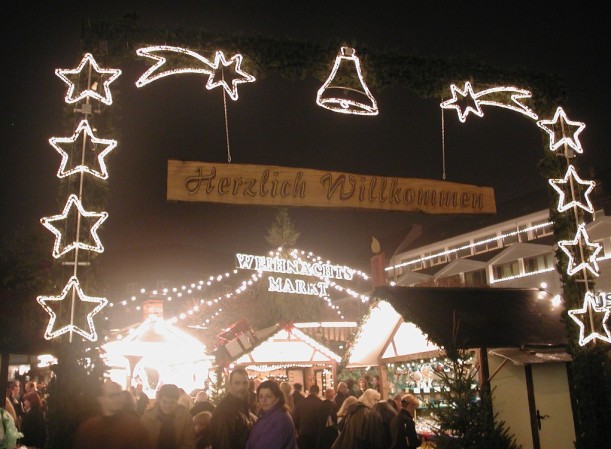 Geschmückter Eingang zum Weihnachtsmarkt. Torähnlich spannt sich ein Bogen aus Tannengrün über den Fußweg. Daran befestigt sind beidseitig mehrere Sterne, mittig ist ein Glöckchen zu erkennen. Die Motive bestehen aus Lichterketten. Unter dem Bogen hängt ein hölzernes Schild mit der Aufschrift "Herzlich Willkommen". Im Hintergrund sind mehrere Verkaufsstände und zahlreiche Weihnachtsmarktbesucher zu sehen.