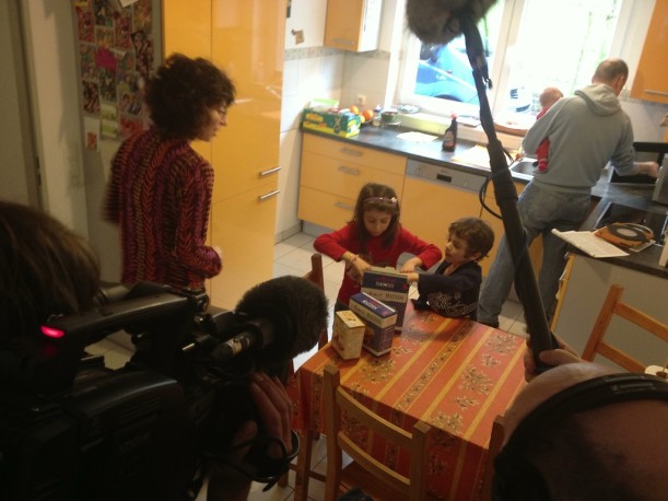 Ein Mann mit Filmkamera und ein Mann mit Tonangel filmen in einer Küche. Eine Frau wendet sich zwei Kindern zu, die eine Schachtel öffnen, die auf dem Küchentisch steht. Im Hintergrund bedient ein Mann mit Baby auf dem Arm eine Mikrowelle.