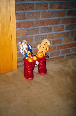 Ein rotes Paar Kinderstiefel steht auf einem braunen Teppich vor einer Backsteinwand. Das Schuhpaar ist mit einem Weckmann, Süßigkeiten wie einem Schokoladennikolaus und Obst gefüllt.