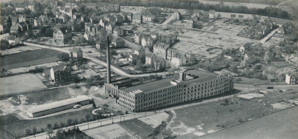 Luftaufnahme der Fabrikgebäude der Schokoladen- und Zuckerwarenfabrik Dr. Hillers AG in Solingen-Gräfrath. Ein Teil des Fabrikgebäudes hat einen sehr hohen Backsteinschornstein. Vor einem anderen Firmengebäude steht ein Firmenlieferwagen. Im Hintergrund sind in lockerer Bebauung Wohngebäude der Gräfrather Ortsteile Ketzberg und Central zu sehen. Das Gelände an den überwiegend neu angelegten Straßen ist noch nicht ganz mit Häusern bebaut; es sind noch viele freie Grundstücke und Gärten vorhanden.