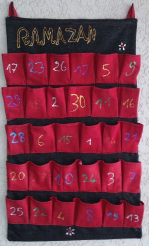 Ramadankalder aus Stoff. Auf einen schwarzen Wandbehang sind 30 rote Täschchen genäht, die jeweils mit Zahlen in unterschiedlichen Farben versehen sind.
