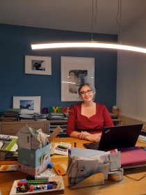 Ein Frau sitze an einem Schreibtisch auf den ein Fülle von Büchern und Büroutensilien leigen, an den Wänden hängen gerahmte Fotos.