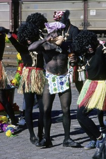 Eine Gruppe als verkleidete Männer stellt sich für den Zug auf. Die Teilnehmer tragen dunkle Lockenperücke, Knochenkette, Baströckchen und dunkle Pullover und Strumpfhosen