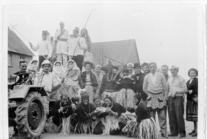 Eine Gruppe ist klischeehaft als schwarze Wilde mit Bastrock und schwarzer Schminke verkleidet, sie posiert vor einem Traktor mit Anhänger. Am Steuer sitzt ein Mann in weißer Uniform im Kolonialstil mit Tropenhut.