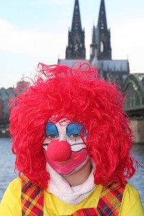 Porträt einer als Clown geschminkten und verkleideten Frau mit roter Schaumstoff-Nase vor dem Rheinpanorama mit Blick auf Kölner Dom und Hohenzollern Brücke.