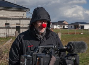 Porträt eines mit einem dunklen Anorak und Kapuze bekleideten Manns, der eine rote Schaumstoff-Nase aufgesetzt hat, in der Hand hält er eine Filmkamera.