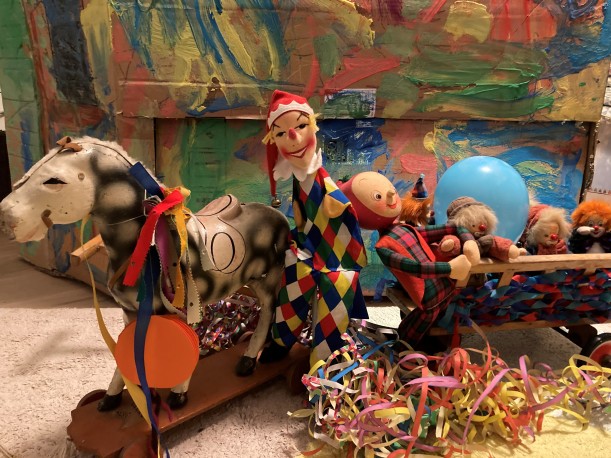 Ein als Karnevalswagen geschmücktes Spielzeug-Wägelchen mit Clowns und einem Hampelmann, das von einem Pferdchen gezogen wird.