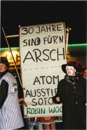 Zentral im Bild ist ein in Druckbuchstaben verfasstes Protestplakat im Hochformat zu sehen. Die Aufschrift lautet: „30 Jahre sind für`n Arsch – Atom Ausstieg Sofort – Robin Wood“. Zwei verkleidete Personen flankieren das Plakat.