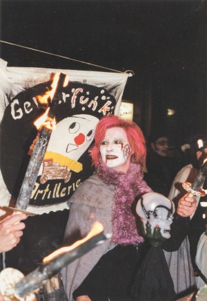 Schaurig kostümierte Frau steht vor einer selbstgemachten Fahne mit der Aufschrift „Geisterfunken Artillerie“. Von links ragen Pechfackeln ins Bild.