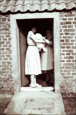 Auf der Schwelle eines Backsteinhauses mit abgesetztem Türrahmen steht eine Frau in hellem, gemustertem Kleid mit einem Säugling auf dem Arm.