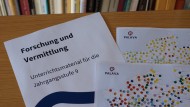 Das Bild zeigt drei Bögen Papier auf einem braunen Tisch. Sie liegen teilweise übereinander. Der Bogen links trägt die Aufschrift "Forschung und Vermittlung. Unterrichtsmaterial für die Jahrgangsstufe 9." Die anderen Bögen sind teilweise sichtbare Sprachkarten der Sprach-App PALAVA.