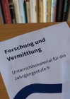 Das Bild zeigt drei Bögen Papier auf einem braunen Tisch. Sie liegen teilweise übereinander. Der Bogen links trägt die Aufschrift "Forschung und Vermittlung. Unterrichtsmaterial für die Jahrgangsstufe 9." Die anderen Bögen sind teilweise sichtbare Sprachkarten der Sprach-App PALAVA.
