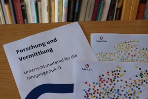 Das Bild zeigt drei Bögen Papier auf einem braunen Tisch. Sie liegen teilweise übereinander. Der Bogen links trägt die Aufschrift "Forschung und Vermittlung. Unterrichtsmaterial für die Jahrgangsstufe 9." Die anderen Bögen sind teilweise sichtbare Sprachkarten der Sprach-App PALAVA.