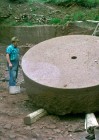 Zwei Männer stehen neben einem großen, steinernen Mühlstein, der auf Holz aufgebockt ist.